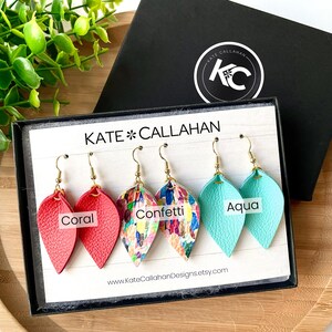 May include: Three pairs of leather earrings in a display box. The earrings are in the colors coral, confetti, and aqua. The box has the text "KATE*CALLAHAN" and a website address.