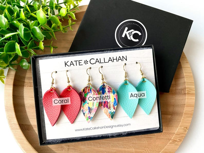 May include: Three pairs of leather earrings in a display box. The earrings are in the colors coral, confetti, and aqua. The box has the text "KATE*CALLAHAN" and a website address.