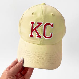 May include: A light yellow baseball cap with the letters "KC" embroidered in red with a white outline. The cap has a curved brim and a button on top. The cap is being held by a person.