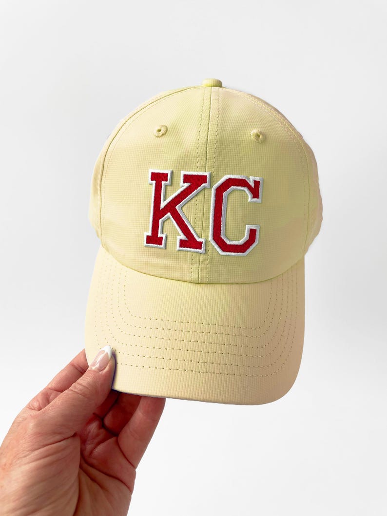 May include: A light yellow baseball cap with the letters "KC" embroidered in red with a white outline. The cap has a curved brim and a button on top. The cap is being held by a person.