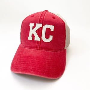 May include: Red and white trucker hat with the letters "KC" embroidered on the front.