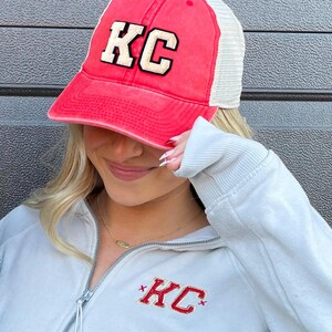 May include: A red and white trucker hat with a gold embroidered "KC" on the front. The hat is being worn by a person wearing a light gray sweatshirt with a red embroidered "KC" on the front.