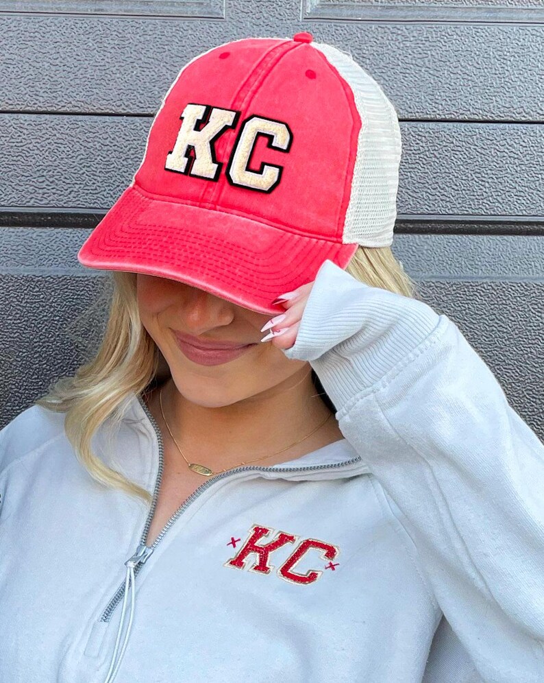 May include: A red and white trucker hat with a gold embroidered "KC" on the front. The hat is being worn by a person wearing a light gray sweatshirt with a red embroidered "KC" on the front.