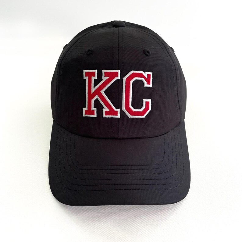 May include: Black baseball cap with the letters "KC" embroidered in red with a white outline on the front. The cap has a curved brim and a structured crown. The hat is made of a durable fabric.