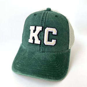 May include: A green and white trucker hat with the letters "KC" in white with a white outline on the front of the hat.