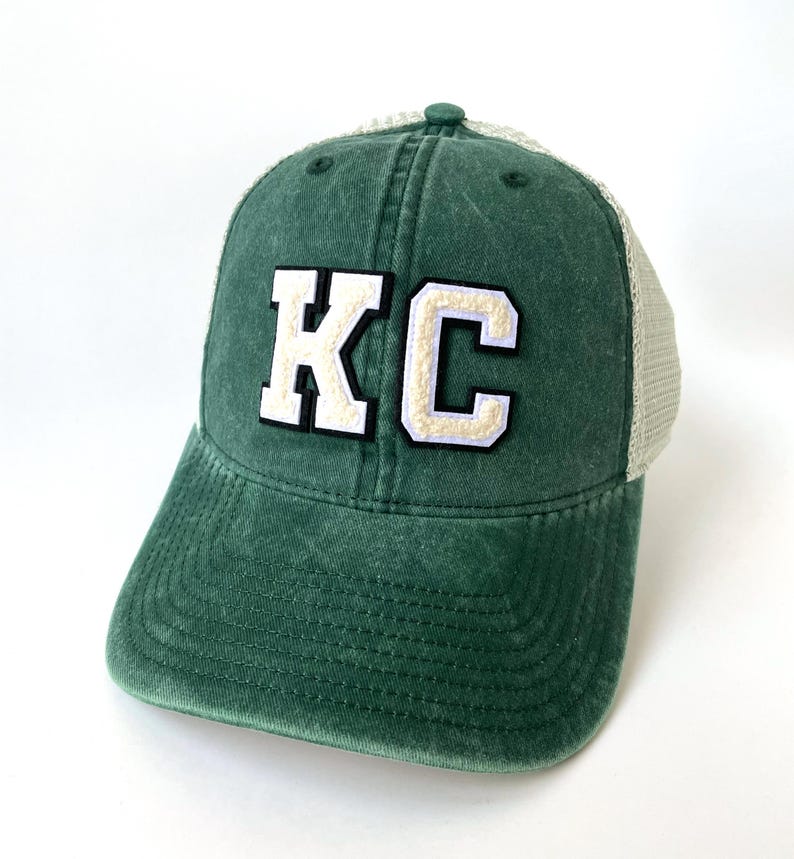 May include: A green and white trucker hat with the letters "KC" in white with a white outline on the front of the hat.