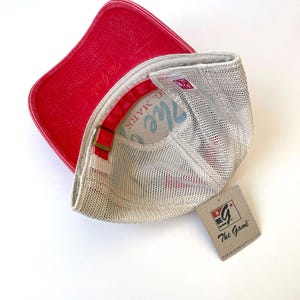 May include: A red and white mesh baseball cap with a red leather peak. The cap has a white mesh back and a red and white logo on the front that says "The Game". The cap is sitting on a white surface.