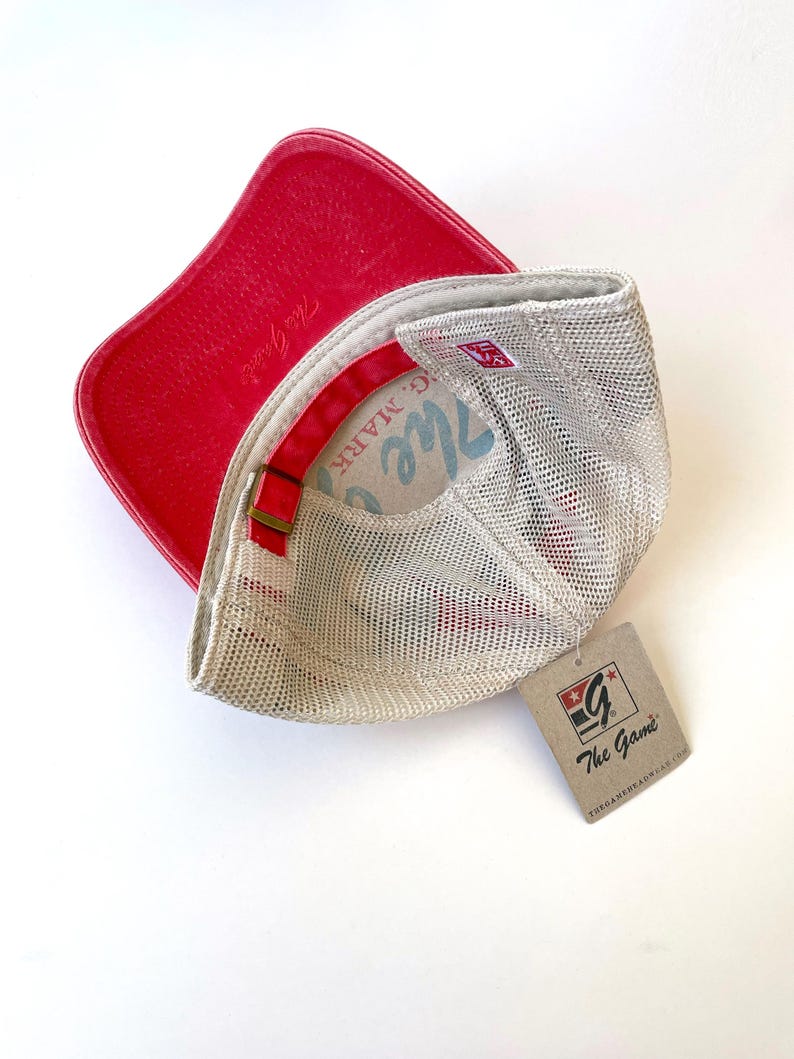 May include: A red and white mesh baseball cap with a red leather peak. The cap has a white mesh back and a red and white logo on the front that says "The Game". The cap is sitting on a white surface.