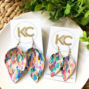 May include: A pair of colorful, teardrop-shaped earrings with a white background and a multi-colored paint splatter design. The earrings are on a white card with the text "Kate Callahan Designs" in gold lettering.