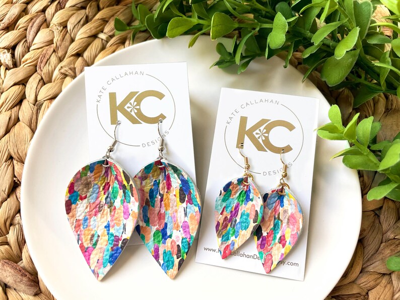 May include: A pair of colorful, teardrop-shaped earrings with a white background and a multi-colored paint splatter design. The earrings are on a white card with the text "Kate Callahan Designs" in gold lettering.