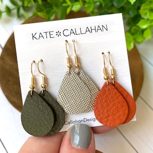 May include: Three pairs of dangle earrings with gold hooks. The earrings are made of leather in green, orange, and a silver-grey color. The earrings are displayed on a white card with the text "KATE*CALLAHAN" and "CallahanDesign".