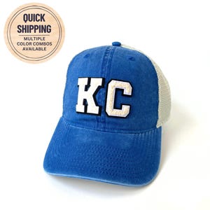 May include: A blue and white trucker hat with the letters "KC" in white with a black outline on the front.