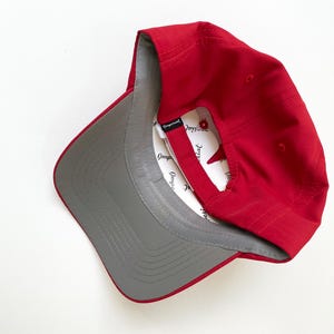 May include: A red baseball cap with a gray underside and a white interior. The cap is angled, showing the interior label. The cap is made of a smooth material.