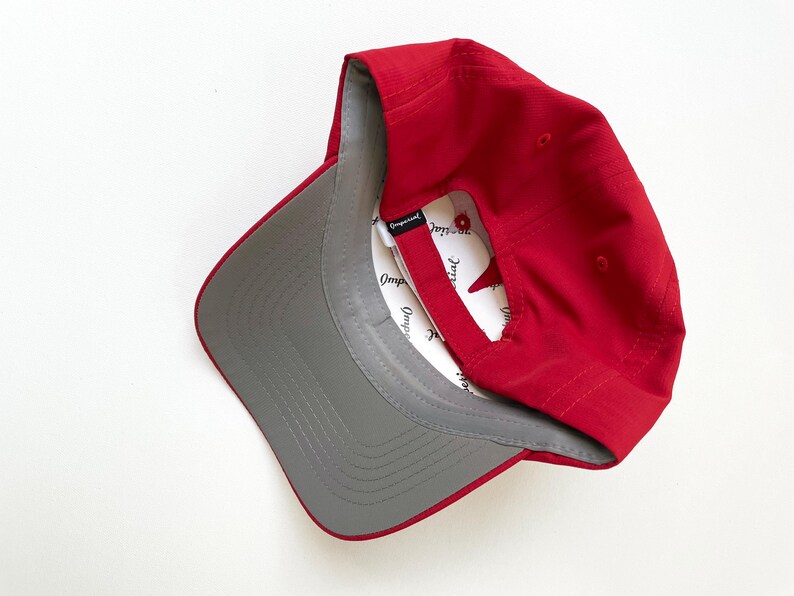 May include: A red baseball cap with a gray underside and a white interior. The cap is angled, showing the interior label. The cap is made of a smooth material.