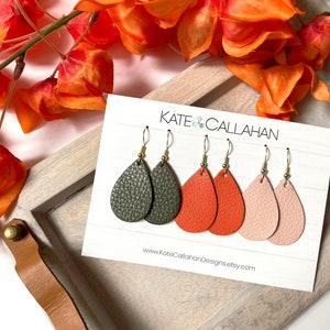 May include: Three pairs of teardrop-shaped earrings on a white display card labeled KATE CALLAHAN. The earrings feature textured faux leather in olive green, burnt orange, and pale pink, each attached to gold-toned metal fishhook ear wires.