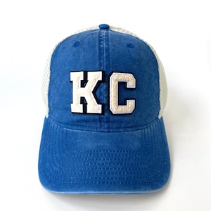 May include: A blue and white trucker hat with a white felt patch that says "KC" in block letters.