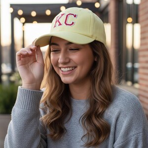 May include: A light yellow baseball cap with the letters "KC" embroidered in red. The cap is worn by a person wearing a gray sweater. The background includes a brick wall and string lights.