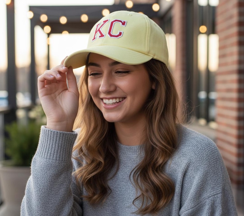 May include: A light yellow baseball cap with the letters "KC" embroidered in red. The cap is worn by a person wearing a gray sweater. The background includes a brick wall and string lights.