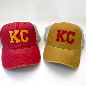 May include: Two trucker hats with mesh back panels. One hat is red with yellow "KC" lettering. The other hat is tan with red "KC" lettering.
