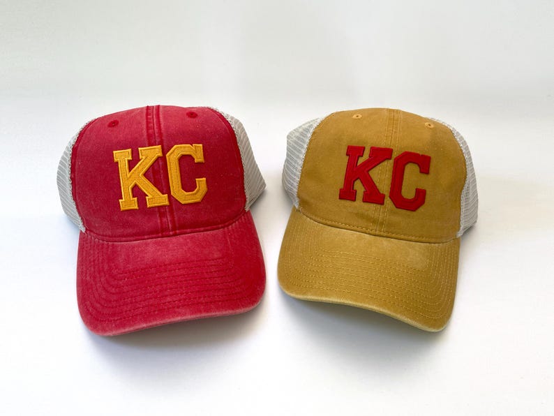 May include: Two trucker hats with mesh back panels. One hat is red with yellow "KC" lettering. The other hat is tan with red "KC" lettering.