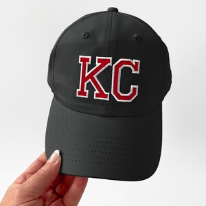 May include: Black baseball cap with the letters "KC" embroidered in red with a white outline. The cap has a curved brim and a textured surface.
