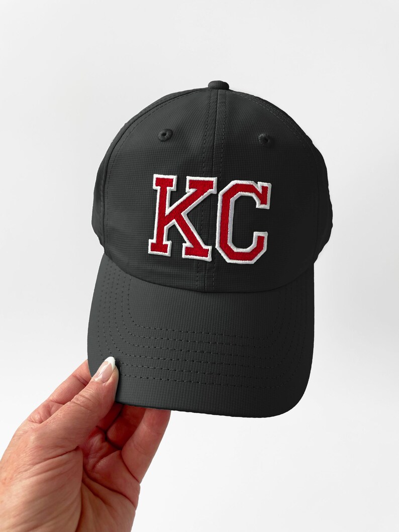 May include: Black baseball cap with the letters "KC" embroidered in red with a white outline. The cap has a curved brim and a textured surface.
