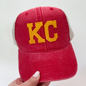 May include: Red and white trucker hat with a yellow embroidered "KC" on the front.