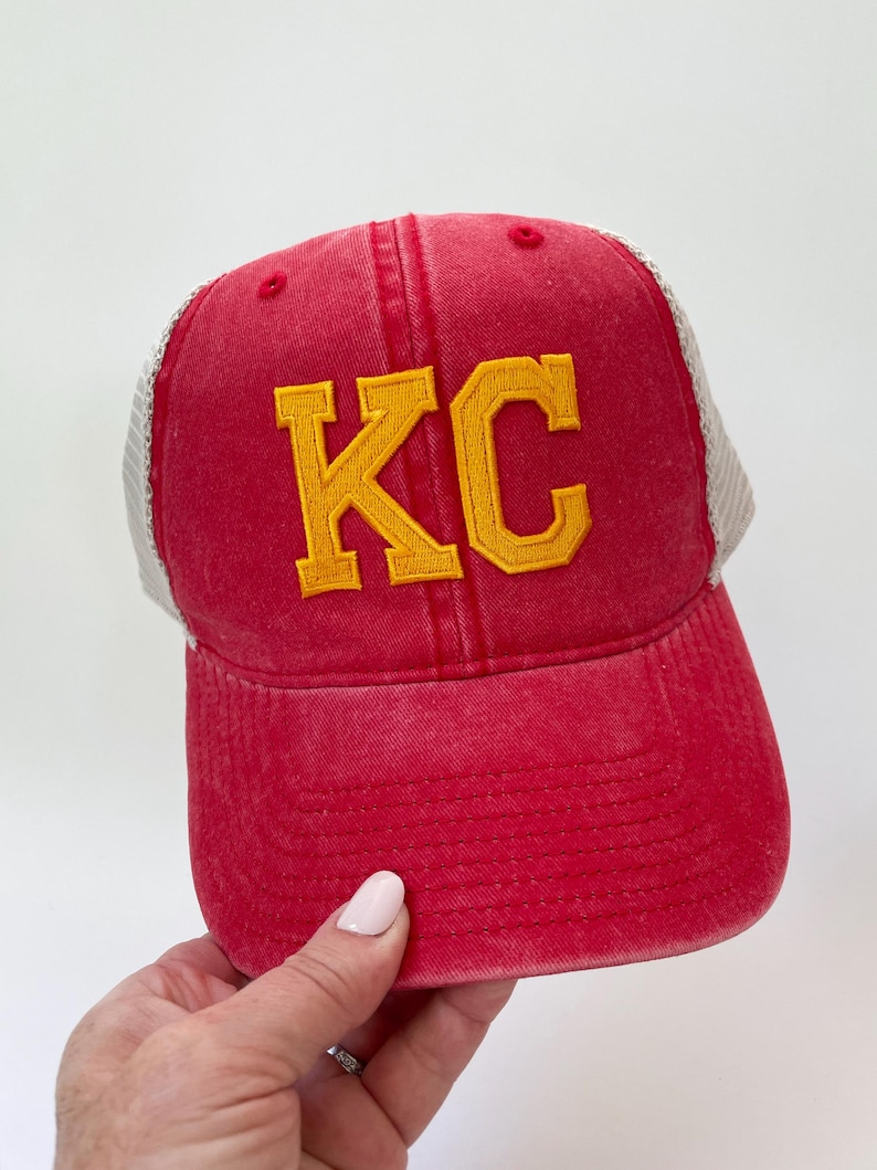 May include: Red and white trucker hat with a yellow embroidered "KC" on the front.