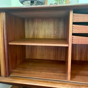 May include: A wooden cabinet with two doors and three drawers. The doors are open, revealing a shelf and empty space. The cabinet is made of a dark wood with a natural finish.