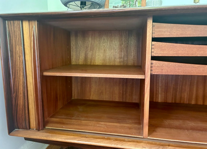 May include: A wooden cabinet with two doors and three drawers. The doors are open, revealing a shelf and empty space. The cabinet is made of a dark wood with a natural finish.