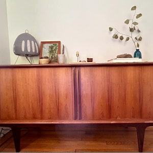 May include: A mid-century modern wooden cabinet with two doors. The cabinet has a light wood finish and is sitting on a wooden floor. There are decorative items on top of the cabinet, including a lamp, a framed picture, and a vase with a branch.