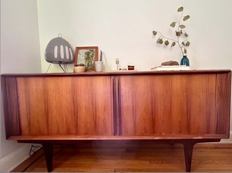 May include: A mid-century modern wooden cabinet with two doors. The cabinet has a light wood finish and is sitting on a wooden floor. There are decorative items on top of the cabinet, including a lamp, a framed picture, and a vase with a branch.