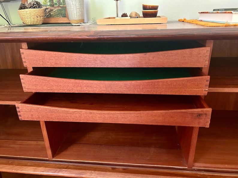 May include: A wooden cabinet with three drawers and open shelves. The drawers are made of a light wood and have a green felt lining. The shelves are made of a darker wood.