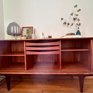 May include: A mid-century modern teak wood sideboard with two sliding doors, three drawers, and open shelves. The sideboard has a rich reddish-brown color and features a unique grain pattern. The sideboard is sitting on a wooden floor.
