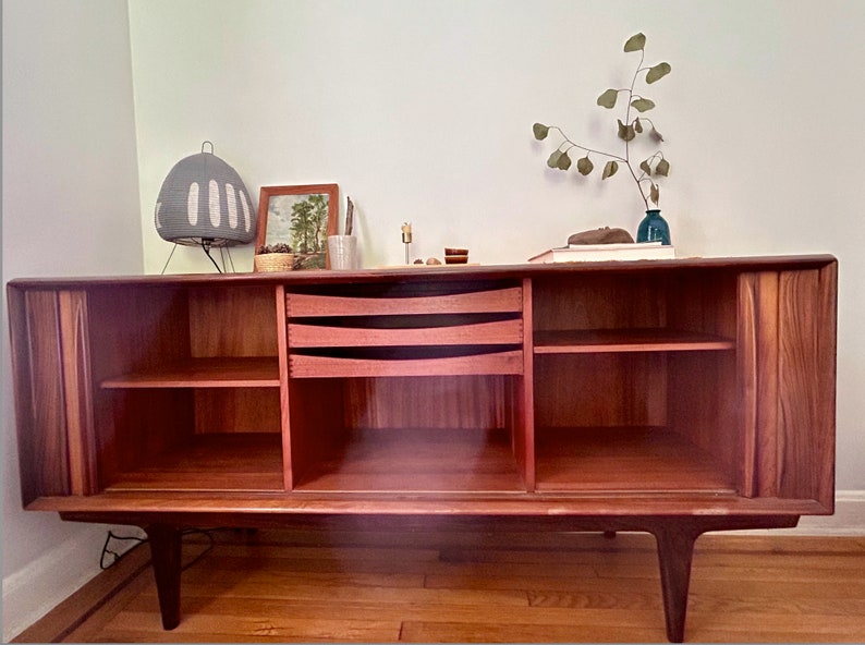 May include: A mid-century modern teak wood sideboard with two sliding doors, three drawers, and open shelves. The sideboard has a rich reddish-brown color and features a unique grain pattern. The sideboard is sitting on a wooden floor.