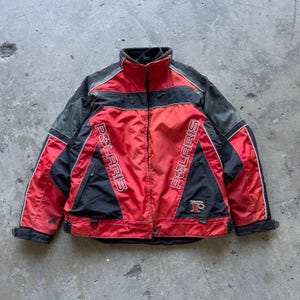 May include: A red and black Polaris jacket featuring a high collar, a full-length front zipper, and long sleeves. The word Polaris is printed vertically in white on both sides of the chest. A small Polaris logo patch is visible on the lower left.