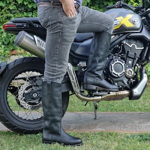 Puede incluir: Una persona con botas de cuero negras con una motocicleta en el fondo. La motocicleta es negra con un tubo de escape plateado.