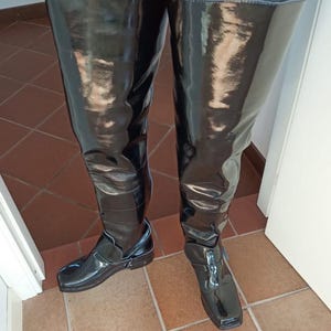 Op de afbeelding: Zwarte, dijhoge laarzen met een glanzende afwerking. De laarzen hebben een vierkante neus en een gespdetail bij de enkel. Gedragen met een blauwe jeans.