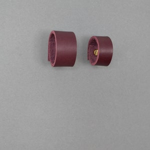 Könnte beinhalten: Zwei burgunderfarbene Lederringe, einer mit einem Messingniet.