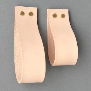 Pode incluir: Dois puxadores de armário de couro rosa claro com parafusos de latão. Os puxadores têm a forma de uma lágrima e uma superfície lisa ligeiramente texturizada.