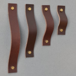 Könnte beinhalten: Fünf braune Leder-Schrankgriffe mit Messing-Schrauben. Die Griffe sind in verschiedenen Größen und haben eine gebogene Form.