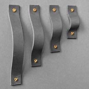 Peut inclure: Quatre poignées de meubles en cuir gris avec des vis en laiton. Les poignées ont un design incurvé et ondulé et sont montées sur un fond gris. Les poignées varient en longueur.