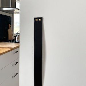 Peut inclure: Poignée de meuble en cuir noir avec des rivets en métal doré. La poignée est fixée à un mur blanc.