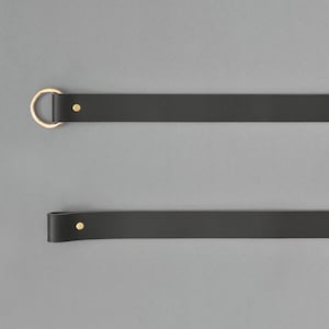 May include: Two black leather straps with gold metal hardware. The top strap has a ring at one end and a small gold dot at the other. The bottom strap has a small gold dot at one end and is rounded at the other.