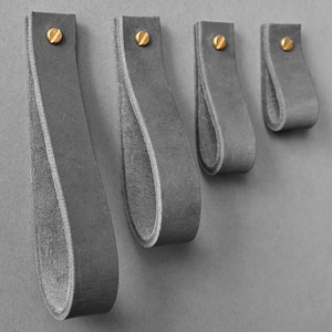 Peut inclure: Quatre poignées de tiroir en cuir gris de différentes tailles sont montées sur un mur gris. Chaque poignée est fixée avec une vis dorée. Les poignées sont en forme de boucle et en cuir doux et texturé. Pour armoires et tiroirs.
