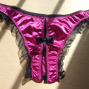 Pode incluir: Roupa interior de cetim rosa com acabamento em renda preta e um laço na frente. A roupa interior tem um recorte nas costas.