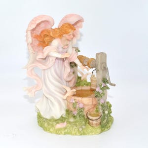 May include: A detailed figurine of an angel with peach-colored wings and flowing red hair, dressed in a white and pink gown. The angel is positioned near a miniature well with a bucket and floral accents, set on a grassy base.