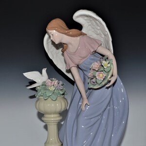 May include: A porcelain figurine of an angel with wings, wearing a pink and blue dress, holding a basket of flowers. The angel is standing next to a white dove perched on a pedestal with flowers.