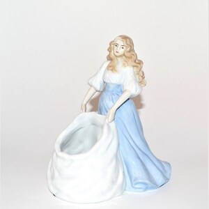 May include: A porcelain figurine of a woman in a blue dress holding a white sack. The woman has long blonde hair and is looking to the side. The sack is open and appears to be a planter.