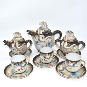 May include: A vintage porcelain tea set featuring a teapot, two sugar bowls, and three teacups with saucers. The set is decorated with a dragon motif in black, gold, and white, with a dragon head on the teapot lid.